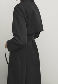 Mustan trench-takki, joka on valmistettu sileästä kankaasta. Ominaisuuksiin kuuluu korkea kaulus, vyötärösolmu ja lyhyt päällysosa, joka ulottuu selän yli.