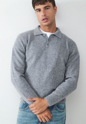 Ung mand med kort krøllet hår iført grå langærmet sweater og blå jeans, stående mod en ensfarvet hvid væg.