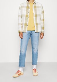 Rutiga skjortan i vitt och oliv, gul T-shirt, ljusblå jeans och krämfärgade sneakers med orange ränder. Modellen står med händerna vid sidorna.