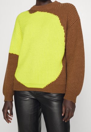 Person iført en brun og neongul strikket sweater med lange ærmer og sorte læderbukser, vist fra skuldre til hofter.