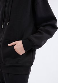 Sudadera negra con cremallera, hecha de un tejido suave, que cuenta con un bolsillo frontal y puños de canalé. Herrajes de cremallera en tono plateado y costuras sin costura.