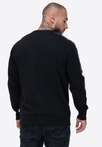 Czarny sweatshirt z długimi rękawami, ribbowym kołnierzem i mankietami. Posiada białe paski po bokach oraz detal z logo w pobliżu dolnej krawędzi. Gładka tekstura materiału.