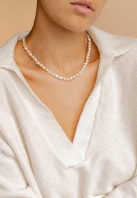 Collar de perlas con cuentas irregulares en forma de óvalo, de un color blanco lustroso, llevado sobre una blusa de tela blanca suave con un diseño de escote en V.