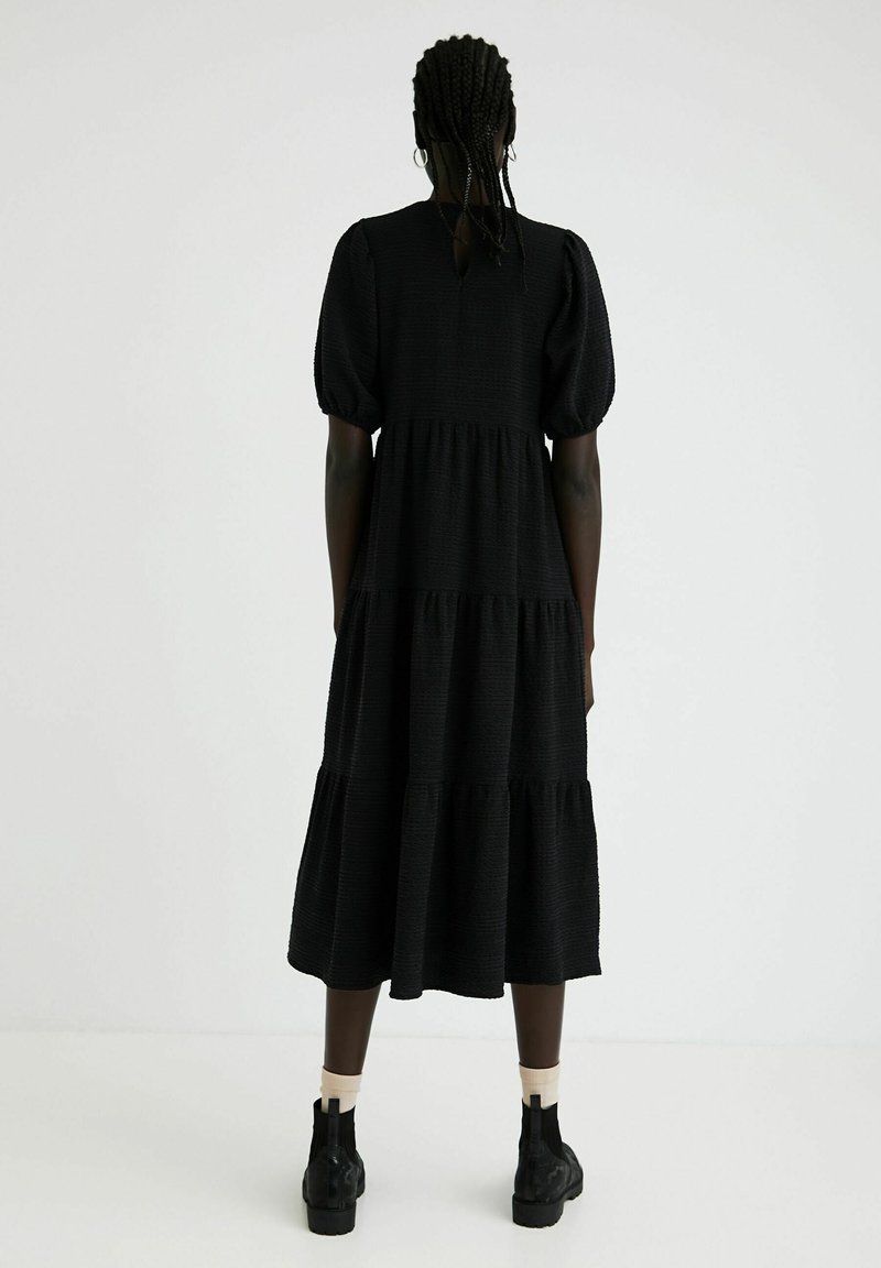 Czarna, warstwowa długa sukienka z bufkami i teksturowanym materiałem. Szczelina w kształcie klucza z tyłu, noszona z czarnymi botkami za kostkę.