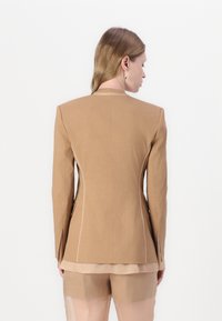 Tan blazer met gestructureerde schouders, contrasterende stiksels en zijsplitten, gecombineerd met een sheer lichtgekleurde onderlaag.