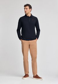 Polo a maniche lunghe blu navy con un logo rosso, abbinato a chinos beige e sneakers marroni. Tessuto liscio, design aderente, stile semplice.