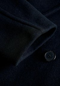 Marineblauwe wollen stof met een zachte textuur, voorzien van een opstaande kraag en een enkel zwart knoopdetail, met een strakke, schone lijnenspel.