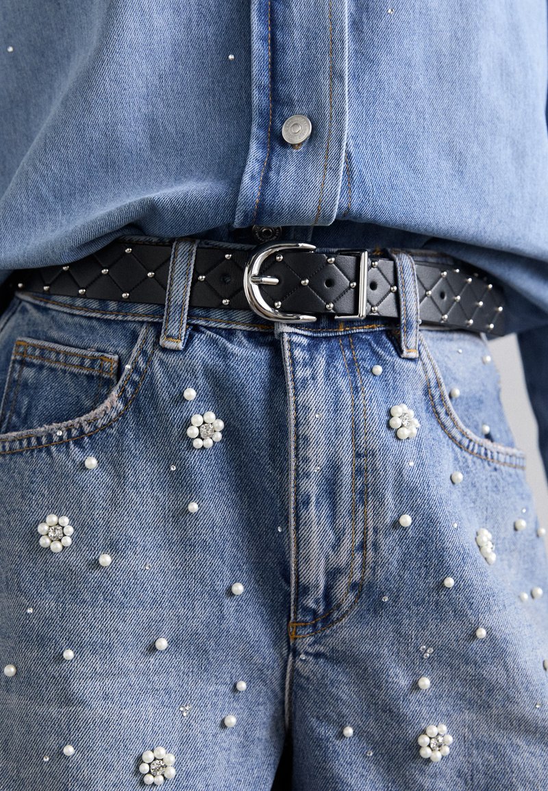 Cintura trapuntata nera con fibbia argentata, indossata sopra jeans in denim chiaro con vari rivestimenti in perle e strass.