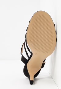 Sandale à talon en daim noir avec un talon étroit et plusieurs straps. La semelle est en caoutchouc beige avec des rainures de traction, indiquant une taille 37.