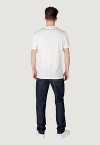 Camiseta de algodón blanca de manga corta, combinada con unos vaqueros de denim azul oscuro y zapatillas blancas, vista desde la parte de atrás.