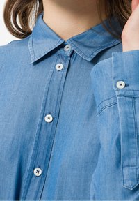 Jasnoniebieska koszula z dżinsu z klasycznym kołnierzykiem, zapięciem na guziki w białym kolorze oraz długimi rękawami zakończonymi mankietami na guziki.