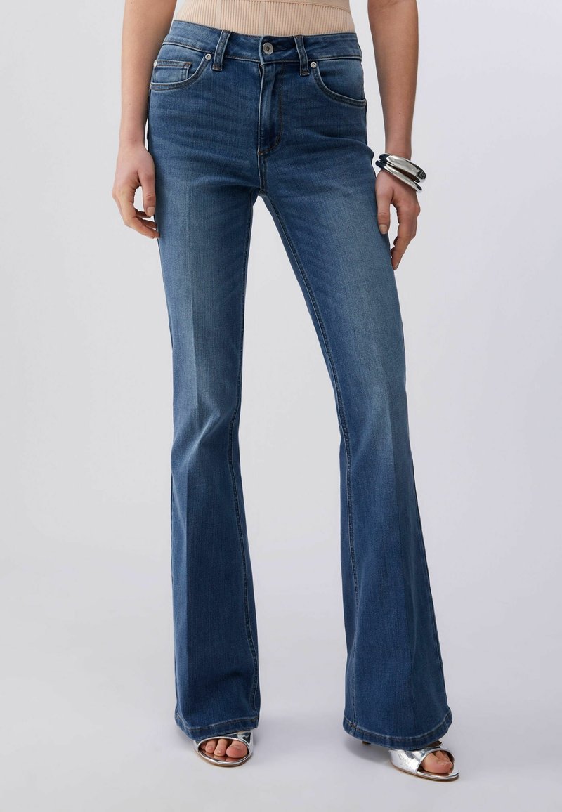 Femme portant un jean flare taille haute bleu, un haut beige, des sandales argentées et des bracelets argentés épais à un poignet, debout devant un fond uni.