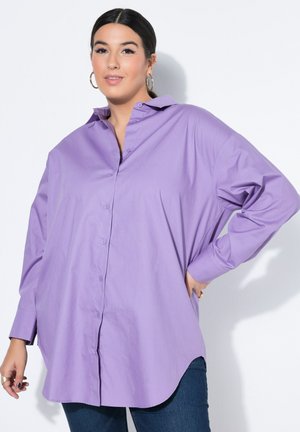 Frau trägt übergroßes langärmeliges lavendelfarbenes Hemd mit Kragen und dunkelblaue Jeans, posiert mit einer Hand an der Hüfte vor weißem Hintergrund.