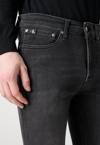Vaqueros de mezclilla negra con un corte ajustado, que presentan herrajes plateados, un zip visible y un parche con el logo de CK encima del bolsillo delantero.