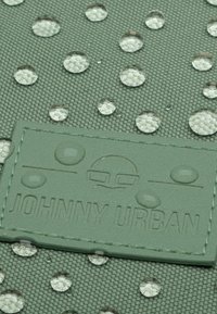 Vízcseppek textúrált zöld szöveten, egy zöld bőr címkével, amelyen a "JOHNNY URBAN" felirat és egy logó látható.