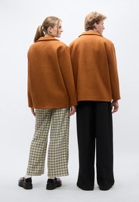 Δύο άτομα στέκονται πλάτη με πλάτη, φορώντας φαρδιές μάλλινες ζακέτες σε σκουριά χρώμα. Ο ένας φοράει καρό παντελόνια με σχέδιο, ο άλλος μαύρο παντελόνι.