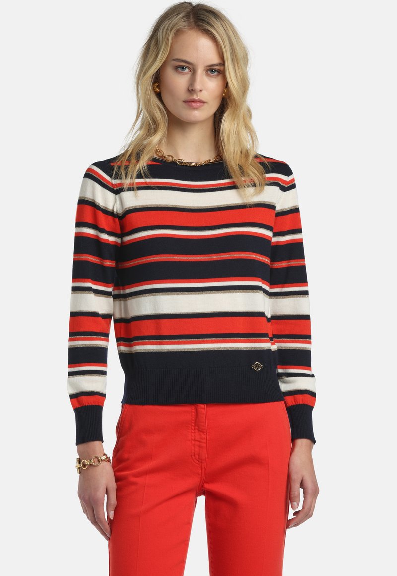 Pull rayé en bleu marine, rouge et crème ; col et ourlet côtelés. Comprend un accent logo doré à la taille. Associé à un pantalon rouge.