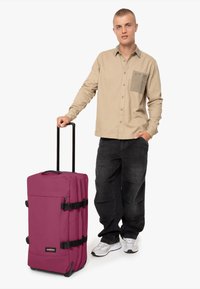 Zaino a rotelle viola con spallacci neri e una maniglia, realizzato in materiale morbido, caratterizzato da una forma strutturata e una maniglia superiore per il trasporto.