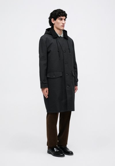 Man standing wearing a black hooded coat, brown pants, and black shoes against a plain light background.