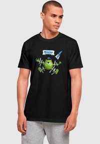 Melna kokvilnas T-krekls ar grafiku, kurā attēlots zaļš varonis no Monsters University, ko ieskauj zili un balti reklāmas uzraksti.