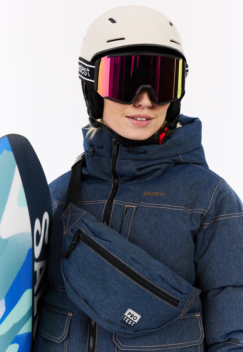 Niebieska kurtka dżinsowa z zamykanymi kieszeniami, czarny kask narciarski, duże lustrzane gogle i niebieska deska snowboardowa z abstrakcyjnymi wzorami. Akcenty w kolorze pomarańczowym.