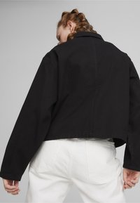 Veste noire coupée avec un col, manches surdimensionnées et coutures visibles. Associée à un pantalon blanc, affichant un design minimaliste.