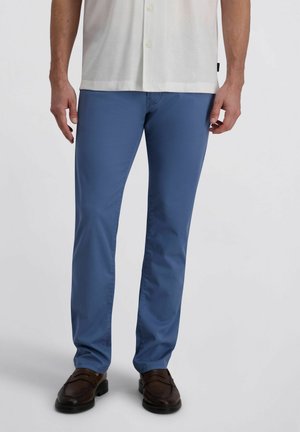 Mężczyzna ubrany w niebieskie, dopasowane spodnie, białą, zapinaną na guziki koszulę oraz ciemnobrązowe skórzane mokasyny, stojący na tle jednolitego tła.