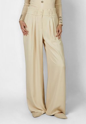 Woman wearing high-waisted, wide-leg beige trousers with pleats, paired with a beige ribbed long-sleeve top and matching pointed shoes.