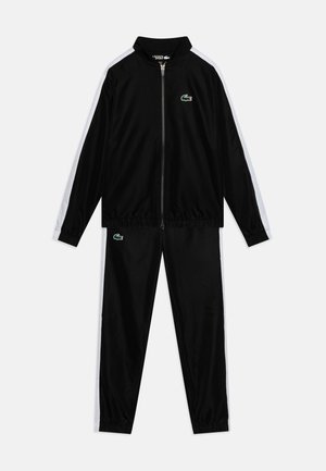Survêtement noir en tissu léger et lisse. Veste avec fermeture éclair, rayures blanches sur les côtés et petit logo vert. Pantalon avec taille élastique.