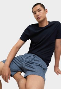 Joven sentado con una pierna doblada, vistiendo una camiseta negra y pantalones cortos azules con estampado sobre un fondo claro y liso.