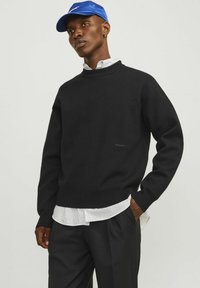 Schwarzer Strickpullover mit geripptem Rundhalsausschnitt, über einem weiß-schwarz gestreiften Hemd getragen, kombiniert mit schwarzen Hosen und einer blauen Mütze.