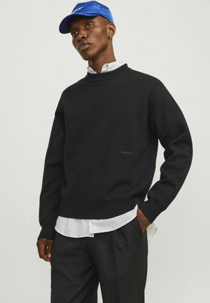 Black knit sweater with a ribbed crew neckline, layered over a white and black striped shirt, paired with black trousers and a blue cap.