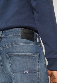 Džínové kalhoty s světle modrým omakem, s černou koženou cedulkou Tommy Jeans na zadním pásku a malým detailem vlajky na kapse.