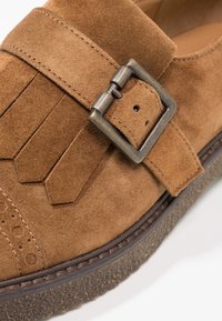 Gros plan sur une chaussure en daim marron avec une sangle décorative, une boucle en métal et une semelle en caoutchouc texturée.