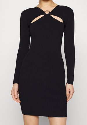 Žena v úzkých černých žebrovaných pletených šatech s dlouhými rukávy a kulatým výřezem na hrudi, který je držán zlatým kroužkem.