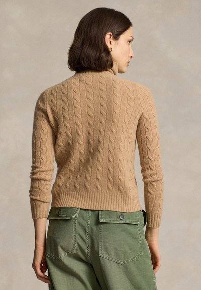 Femme vue de dos portant un pull à col roulé torsadé couleur chameau et un pantalon cargo vert olive sur un fond neutre.