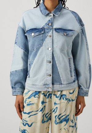 Osoba ubrana w patchworkową kurtkę jeansową w odcieniach jasnoniebieskiego i ciemnoniebieskiego z metalowymi guzikami oraz kremowe spodnie z niebieskimi abstrakcyjnymi wzorami.