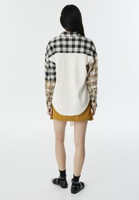 Camisa de cuadros con un panel blanco texturizado en la parte posterior, con mangas largas y dobladillo redondeado. Lleva puesta una falda amarilla y zapatos negros.