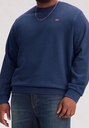 Man draagt een marineblauwe Levi's sweatshirt met een klein rood logo op de borst en donkerblauwe jeans, tegen een effen achtergrond.