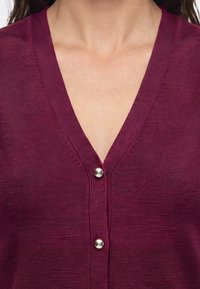 Femme portant un cardigan bordeaux à col en V avec deux boutons ronds argentés fermés à l'avant.