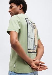 Kaaslastega mees, kes kannab helerohelist T-särki abstraktse ristkülikukujulise mustriga seljal, käed selja taga kokkukõrvatud, hele sinised teksad.