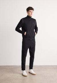 Trening negru cu fermoar, fabricat dintr-un material moale, cu guler înalt și dungi pe lateral, asortat cu adidași albi cu talpă din cauciuc.