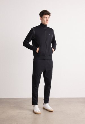 Zwarte trainingsoverall met rits, gemaakt van zachte stof, met een hoge kraag en zijstrepen, gecombineerd met witte sneakers met gumzolen.