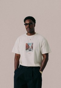 Beige T-Shirt mit kurzen Ärmeln, das ein Vorderdesign einer Stadtlandschaft zeigt, kombiniert mit dunklen Hosen. Das Shirt hat eine lockere Passform und einen runden Halsausschnitt.