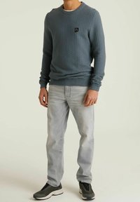 Blue textured sweatshirt with a round neck and small logo on the chest, paired with light grey jeans and black sneakers.