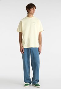 Hellgelbes Baumwoll-T-Shirt mit Rundhalsausschnitt und kurzen Ärmeln, kombiniert mit blauen lässigen Jeans und grünen Sneakern. Kleines Logodetail.