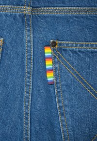 Blå denimstoff med oransje sømmer, og en flerfargetstripet dekorative lapp på baklommen. Ingen synlige metalldetaljer.