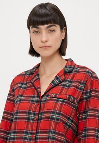 Rød ternet skjorte med knaplukning, der har sort piping, en brystlomme og en afslappet pasform med krave og lange ærmer.