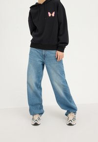 Sudadera con capucha negra con un gráfico de mariposa rosa; combinada con jeans azules sueltos y zapatillas de color claro con un patrón de olas.