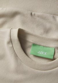 Beige crewneck sweatshirt with ribbed collar and a green fabric label reading "JJXX" sewn inside the neckline.